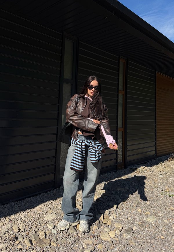 Femme portant des lunettes de soleil, une veste en cuir marron, une chemise rose, un jean et un pull rayé noué autour de la taille, debout sur un sol rocheux près d’un bâtiment sombre.