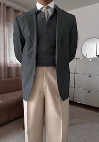 Homme portant un blazer gris, un pull gris foncé par-dessus une chemise blanche avec une cravate rayée, et un pantalon beige, debout à l'intérieur près d'un canapé et d'un buffet.
