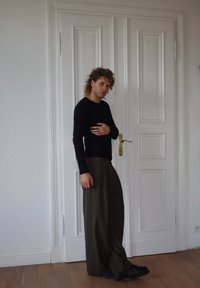 Top nero a maniche lunghe, pantaloni larghi marroni a righe verticali e scarpe nere. In piedi accanto a una porta bianca a pannelli in una stanza ben illuminata.