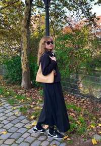 Jupe noire et veste noire raccourcie, sac beige et baskets noires. La mannequin a de longs cheveux ondulés et porte des lunettes de soleil sur fond naturel.