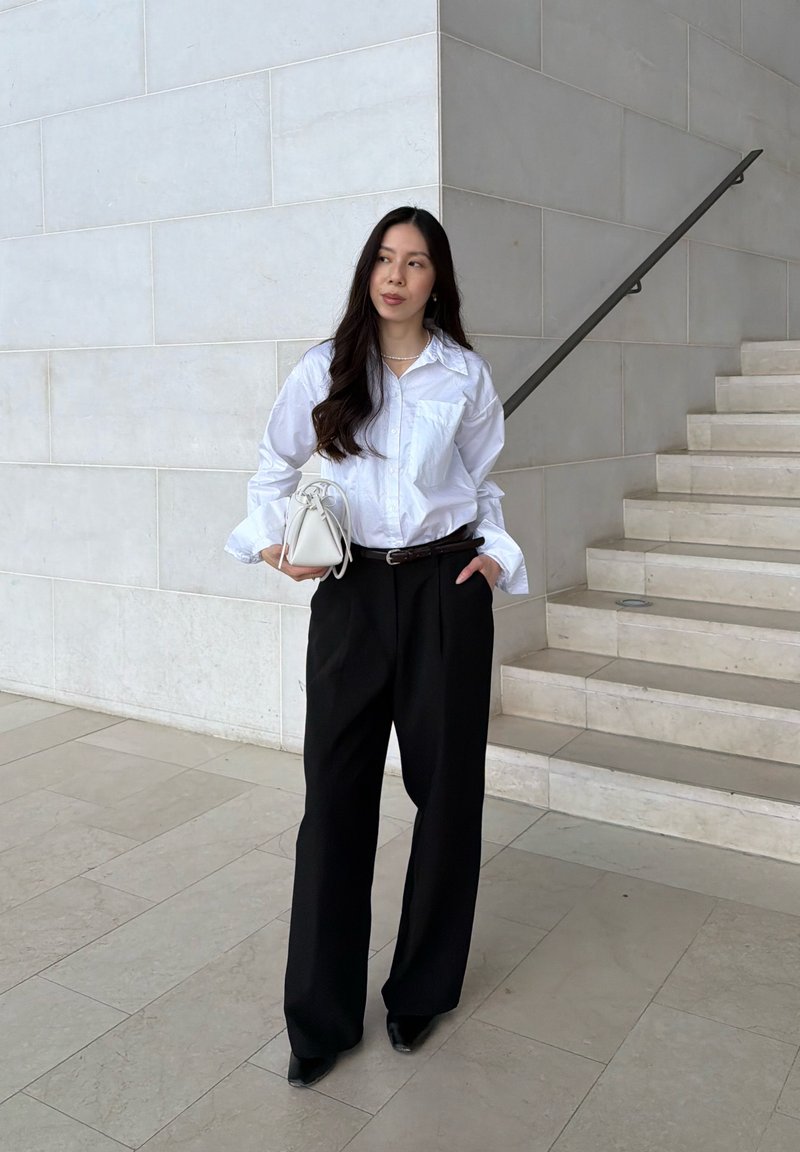 Donna con capelli lunghi e scuri, indossa una camicia bianca con bottoni e pantaloni neri a gamba larga, tiene una borsa bianca vicino alle scale.