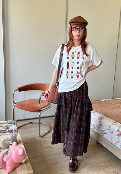 Persona joven que lleva un sombrero marrón, gafas, camiseta blanca con estampado de reloj, falda larga de cuadros y zapatos marrones, de pie en una habitación con silla y cama.