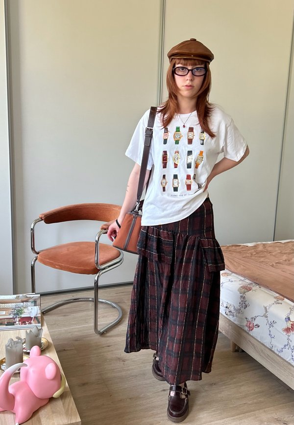 Persona joven que lleva un sombrero marrón, gafas, camiseta blanca con estampado de reloj, falda larga de cuadros y zapatos marrones, de pie en una habitación con silla y cama.