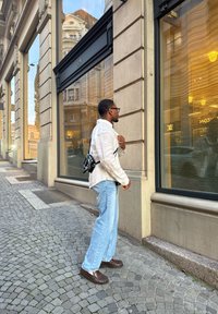 Inimene valges linases särgis ja helesinistes teksapükstes seisab poeakna kõrval, kehast üle musta koti rippumas. Munakivitee.