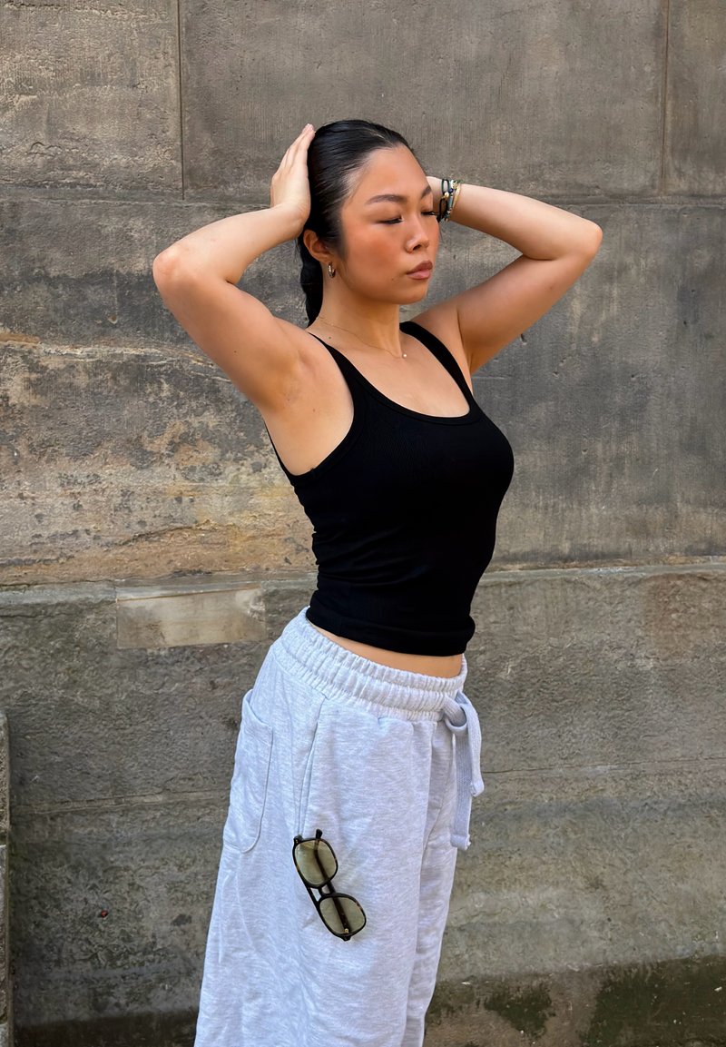 Femme en débardeur noir et pantalon de jogging gris se tient contre un mur en pierre, les mains derrière la tête et des lunettes de soleil suspendues à la poche.