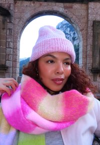 Femme aux cheveux bouclés portant un bonnet en tricot rose et une écharpe colorée rose-jaune, debout en plein air avec une arche en pierre et une montagne en arrière-plan.