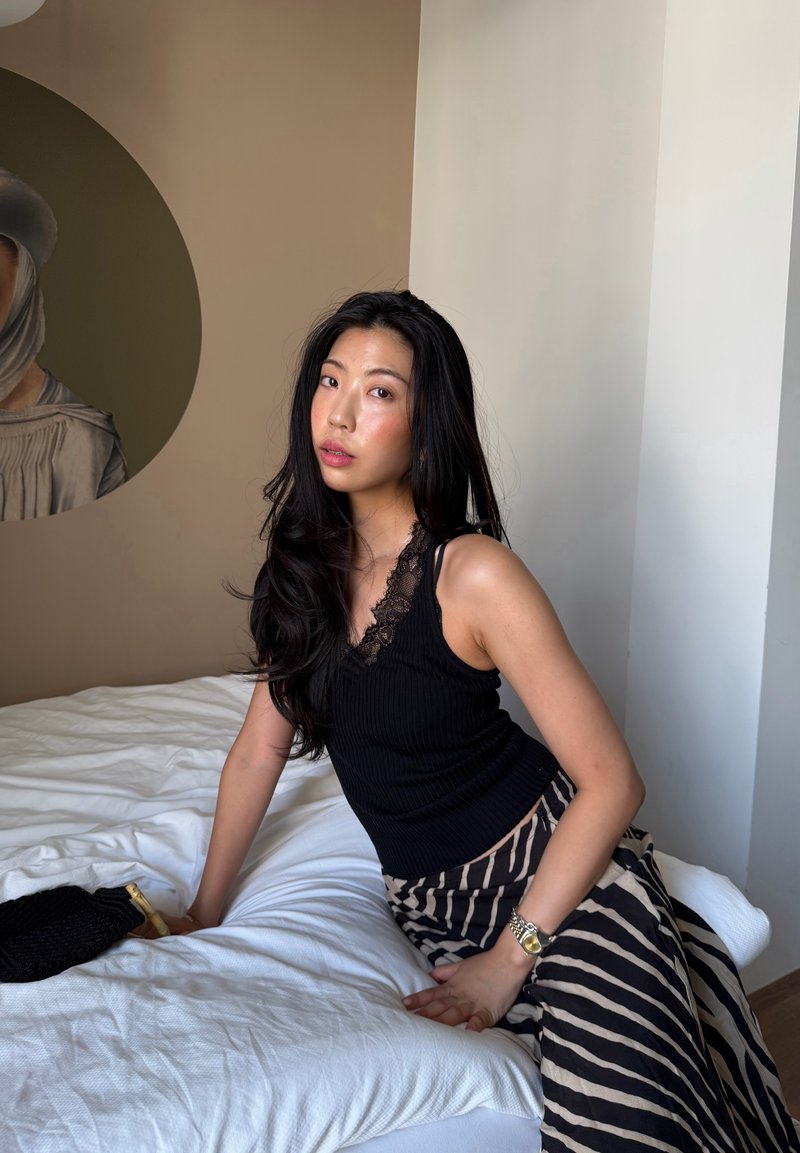 Femme aux longs cheveux noirs portant un haut noir et une jupe à rayures, assise sur un lit avec des draps blancs dans une pièce aux tons neutres.