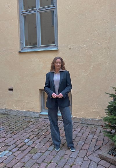 Blazer negro ajustado sobre una blusa de punto clara, combinado con pantalones de pierna ancha grises y zapatos casuales, de pie sobre adoquines cerca de una pared amarilla.