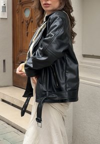 Fekete bőr motoros dzseki laza fazonnal, cipzáras zsebekkel és övvel ellátott alsó résszel. A felülete sima textúrájú és enyhe fényességű.