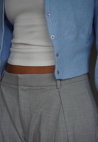 Helles blaues Strickjacke. Weisses, figurbetontes Tanktop darunter. Graue, maßgeschneiderte Hose mit Falten. Der Stoff scheint weich und strukturiert zu sein. Silberne Knöpfe.