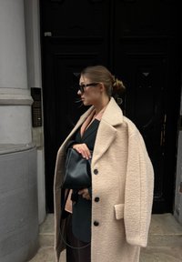 Femme avec les cheveux en chignon portant un manteau beige sur un blazer foncé et des lunettes de soleil, tenant un sac à main noir, debout près d'une porte en bois foncé.