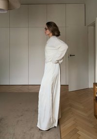 Biały puszysty kardigan w połączeniu z długą białą spódnicą. Pokój z podłogą z jasnego drewna i neutralnymi ścianami. Minimalistyczne elementy wystroju.