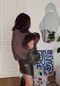 Μαλλινό πουλόβερ σε ήσυχο λεβάντα, μαύρη δερμάτινη μίνι φούστα, πράσινη τσάντα και ένα καφέ σκύλο. Στο φόντο υπάρχουν αφηρημένη τέχνη και περιοδικά.