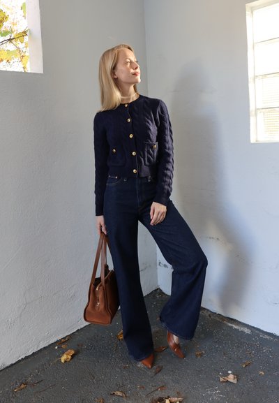 Cárdigan azul marino de punto con botones dorados, combinado con jeans de mezclilla oscura y pierna ancha. El conjunto se completa con un bolso marrón y tacones marrones de punta afilada.