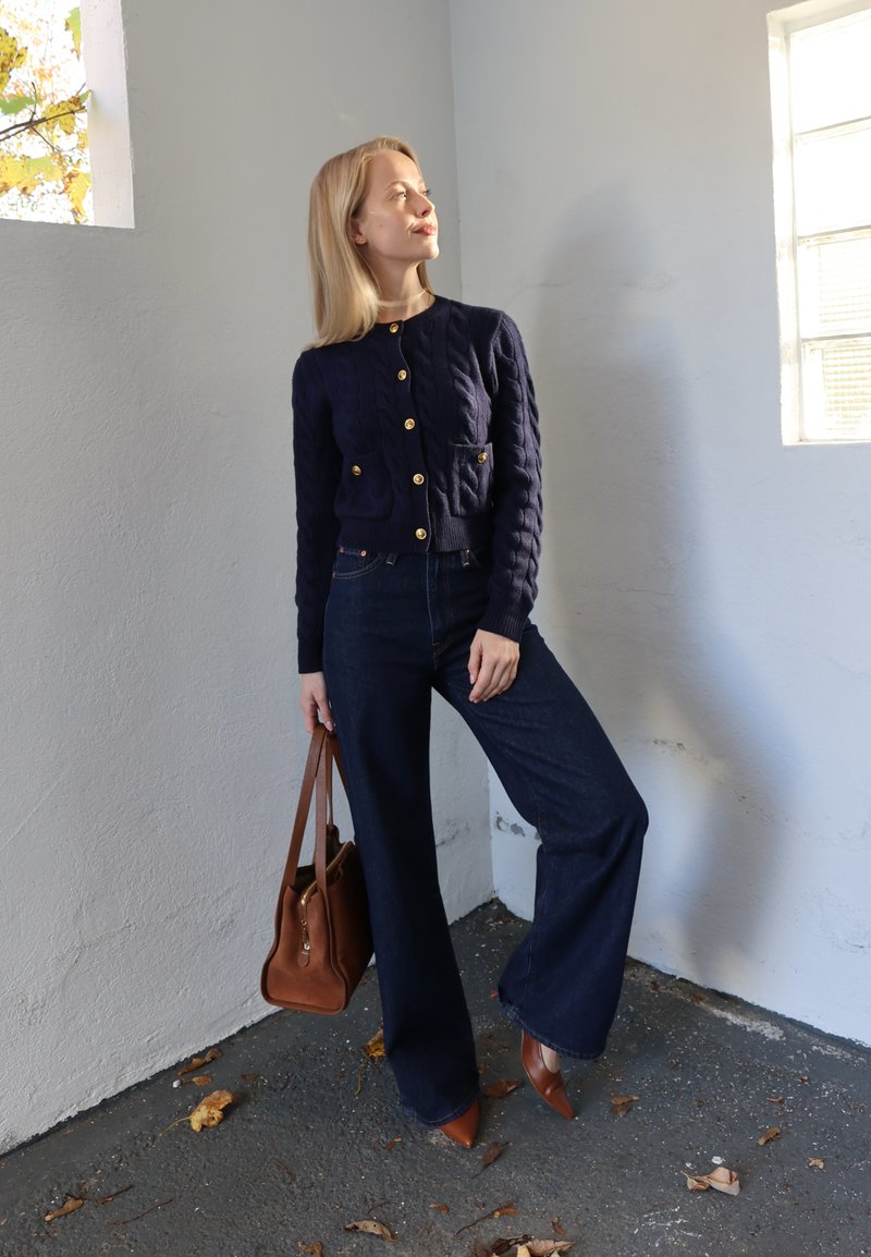 Marineblå cardigan med kabelstrik og guldknapper, kombineret med mørk denim jeans med vide ben. Brun håndtaske og brune spidse hæle fuldender looket.