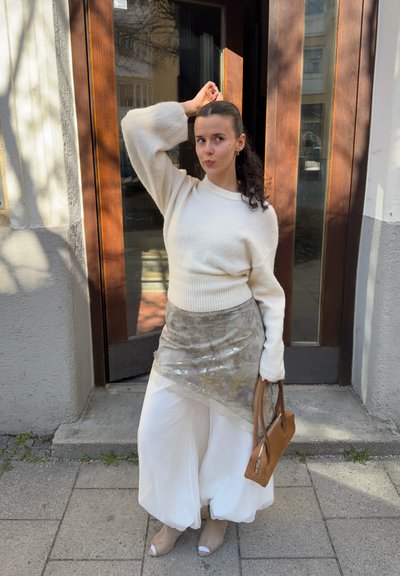 Mujer con cabello rizado oscuro, suéter blanco, falda transparente con estampado sobre pantalones blancos, zapatos de tacón abiertos color marrón claro, sosteniendo un bolso marrón, de pie junto a una puerta de madera.