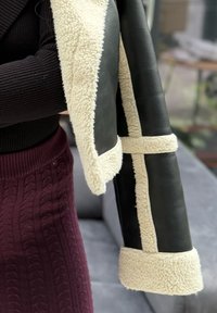 Personne tenant un manteau noir doublé de shearling crème, portant un haut côtelé noir et une jupe tricotée bordeaux.