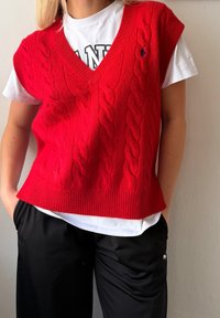Chasuble en maille torsadée rouge avec un col en V, présentant un ourlet côtelé. Portée sur un T-shirt blanc, associée à un pantalon noir.