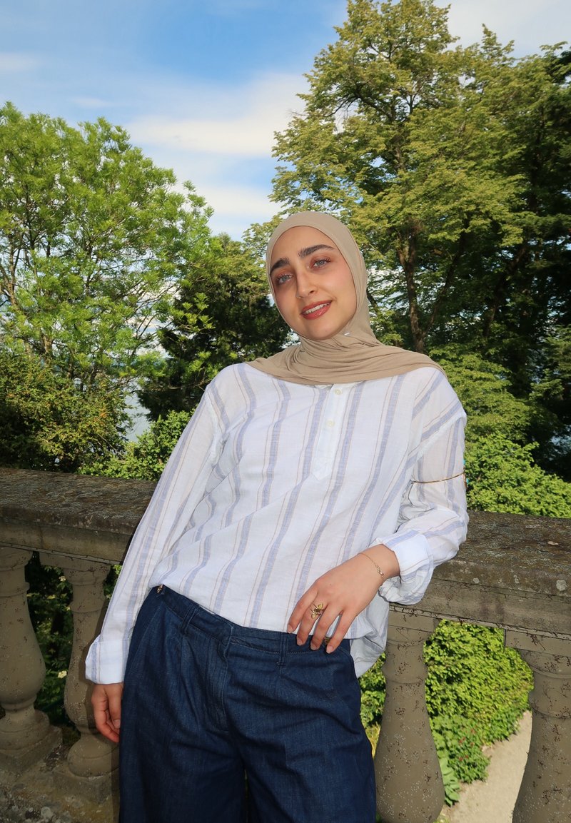 Junge Frau mit beigem Kopftuch und weiß gestreiftem Hemd lehnt an einem steinernen Geländer, im Hintergrund grüne Bäume und blauer Himmel.