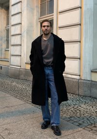 Juodas ilgas paltas virš pilko riboto megztinio, derinamas su šviesiai mėlynais džinsais ir juodais batais. Stovi ant akmeninės grindinio dangos netoli pastato.