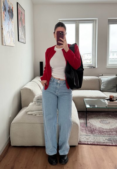 Cardigan rojo, blusa blanca y jeans de pierna ancha de color azul claro. Zapatos negros gruesos y un gran bolso negro. Fondo de sala de estar en tonos neutros.