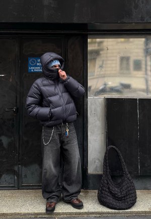 Chaqueta negra acolchada con cuello alto, pantalones vaqueros gris de pierna ancha, zapatos marrones y un bolso negro texturizado de hombro. Cadena unida a los vaqueros.