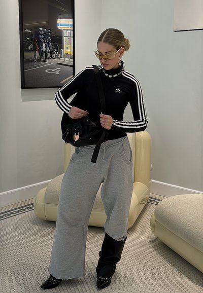 Camiseta negra de Adidas de manga larga con franjas blancas, pantalones de chándal grises y botas negras. Bolso negro con detalles de hardware. Ambiente minimalista.