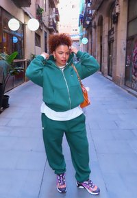 Grønn hettejakke med glidelås og matchende joggebukser med et lag hvit skjorte under. Fargerike mønstrede joggesko og brun skulderveske. Urbant miljø.