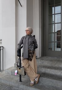 Femme en pantalon beige et haut gris avec écharpe et gants, appuyée sur une rambarde près des marches d'un bâtiment, tenant un sac à main à motifs avec un porte-clés en peluche.