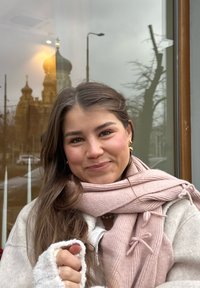 Jeune femme aux longs cheveux bruns portant une écharpe rose et un pull blanc, souriant en plein air devant une fenêtre en verre reflétant un bâtiment et un réverbère.