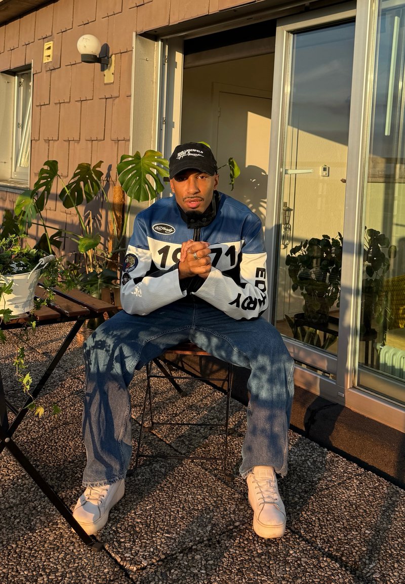 Une personne est assise sur une chaise en métal, portant une veste bleue et blanche avec "1971" et un jean. La lumière du soleil met en valeur des plantes d'intérieur et un cadre de porte.