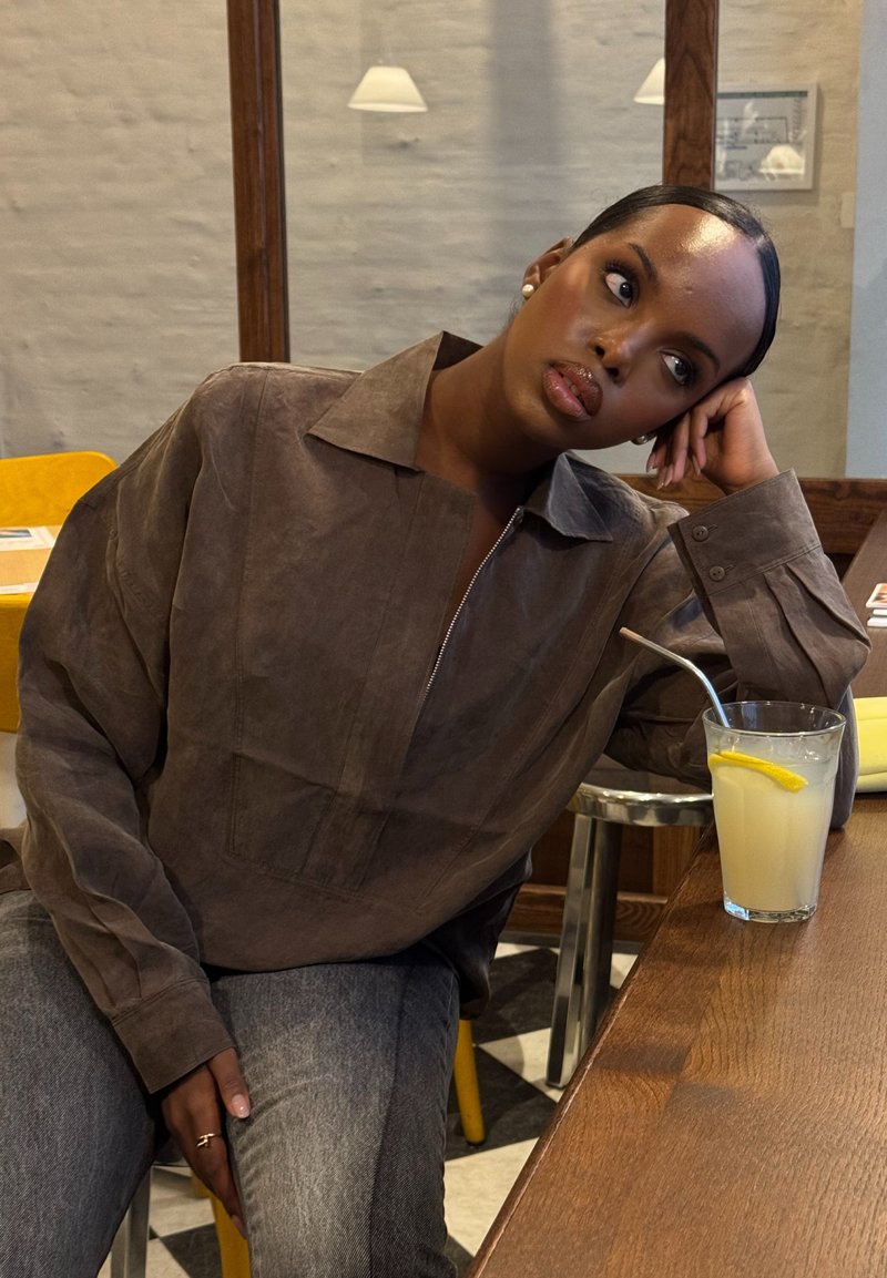 Woman wearing a brown shirt and grey jeans, leaning against a wooden table with a glass of lemonade and a metal straw, gazing thoughtfully to the side.