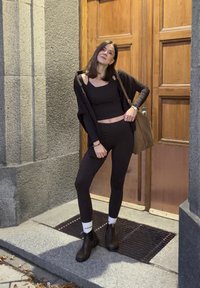 Crna odjeća koja se sastoji od uskog topa i tajica s dugim, otvorenim kardiganom. Smeša torba i gležnjače. Pozadina od drvenih vrata.