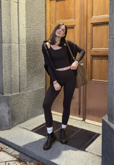 Conjunto negro que incluye una blusa ceñida y leggins, acompañados de un cárdigan largo y abierto. Bolso marrón y botas de tobillo. Fondo de puerta de madera.