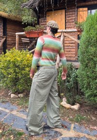 Persona, kas valkā daudzkrāsainu, ribotu džemperi zaļā, sarkanā un brūnā krāsā, kopā ar vaļīgām olīvu zaļām cargo biksēm un melniem apaviem.