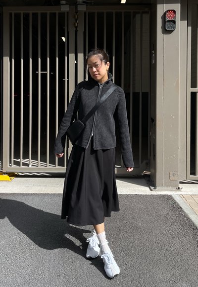 Chaqueta negra de cremallera acanalada, falda midi negra, zapatillas blancas con cordones y calcetines de tobillo, de pie frente a una entrada con rejas.