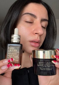 Zwei Estee Lauder Hautpflegeprodukte: eine dunkelbraune Glasflasche mit einem Tropfer, beschriftet mit "Advanced Night Repair", und ein schwarzes Glas mit goldenem Deckel, beschriftet mit "Revitalizing Supreme+."