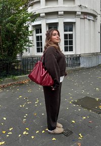 Καφέ κοντό sweatshirt και ταιριαστά παντελόνια, σε συνδυασμό με μπεζ παντόφλες. Μια φωτεινή κόκκινη τσάντα είναι κρεμασμένη στον ένα ώμο. Πλευρική θέα σε αστικό περιβάλλον.