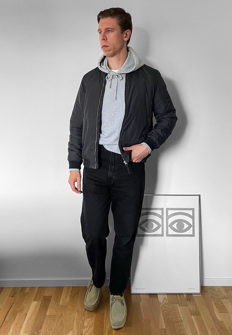 Hombre con chaqueta bomber negra sobre una sudadera con capucha gris, jeans negros y zapatos beige. De pie junto a una obra de arte gráfica enmarcada sobre un suelo de madera.