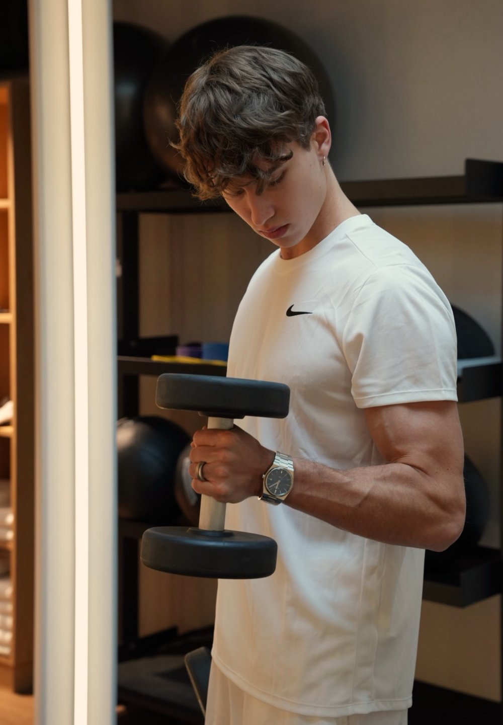 Svart manual med sølvfarget håndtak holdt i én hånd, iført en hvit Nike T-skjorte. Lys bakgrunn med treningsutstyr synlig.