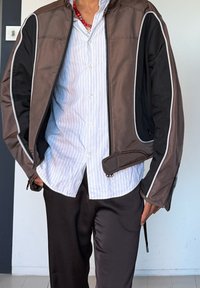 Άτομο που φοράει καφέ και μαύρο μπουφάν με φερμουάρ πάνω από λευκό πουκάμισο με ρίγες, σκούρο παντελόνι και κόκκινο κολιέ με χάντρες.