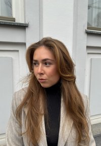 Una mujer con cabello castaño largo y ondulado lleva un blazer de rayas gris claro sobre un suéter de cuello alto negro, frente a una pared blanca.