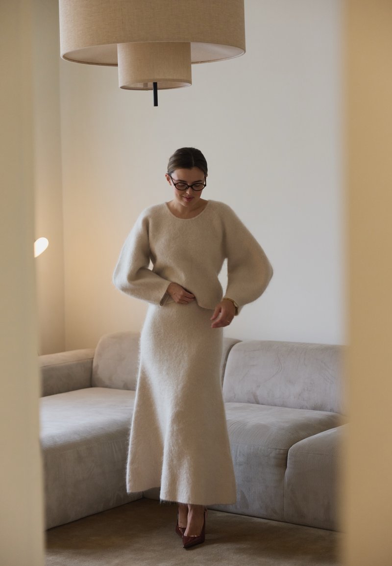 Pull en tricot texturé beige avec des manches volumineuses, associé à une jupe longue assortie, dans un espace de vie doux aux tons neutres.