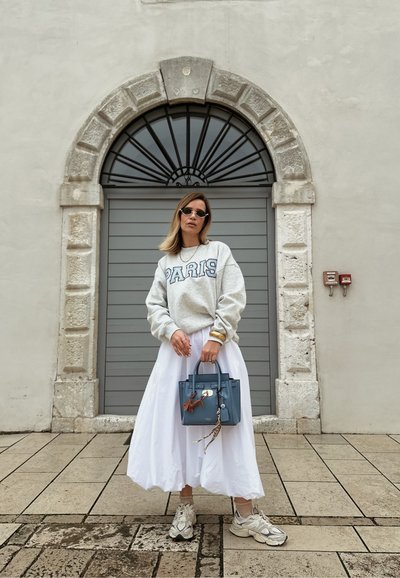 Sudadera gris clara con el texto "PARIS", falda midi blanca, bolso azul y zapatillas ligeras, todo ello delante de una pared de piedra texturizada y una ventana arqueada.