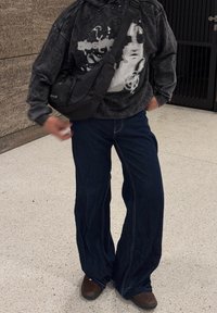 Felpa grafica nera con una vestibilità ampia, abbinata a jeans scuri a gamba larga e scarpe marroni, completata da una borsa a tracolla nera.