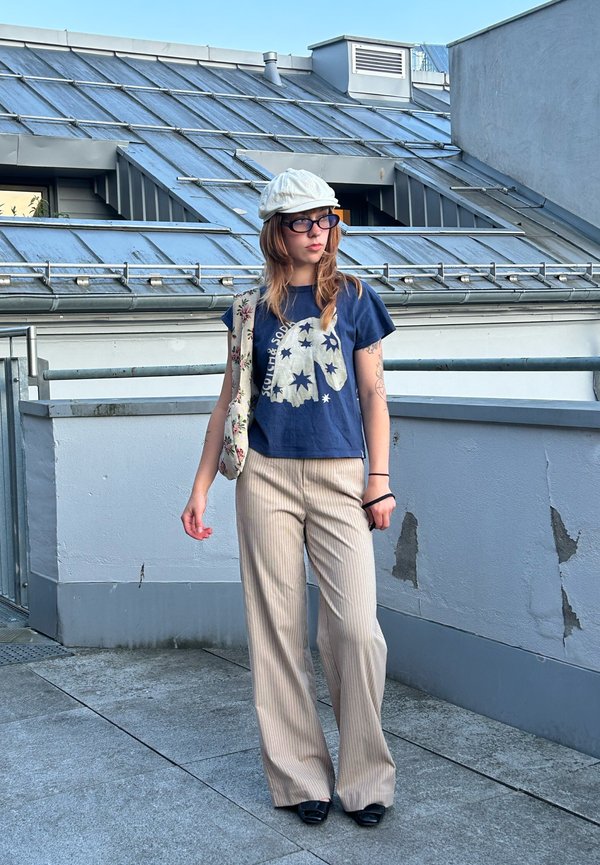 Joven mujer con gorra blanca, gafas, camiseta azul con estampado de estrellas, pantalones beige de rayas y zapatos negros, de pie en una terraza en la azotea con barandilla metálica.