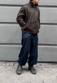 Personne debout contre un mur gris, portant une veste en cuir marron, un pantalon ample bleu foncé et des bottines grises, les mains dans les poches.
