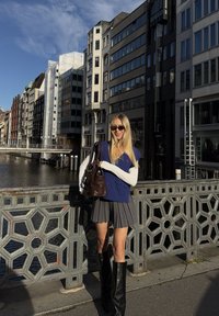 Uma mulher está de pé numa ponte, vestindo um colete de malha azul sobre uma camisola de mangas compridas branca, uma saia plissada cinza e botas pretas até ao joelho.