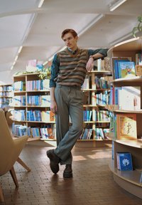 Gilet lavorato a maglia con strisce multicolori sopra una camicia scura con bottoni, abbinato a pantaloni chiari e scarpe scure, in piedi accanto a scaffali di libri.