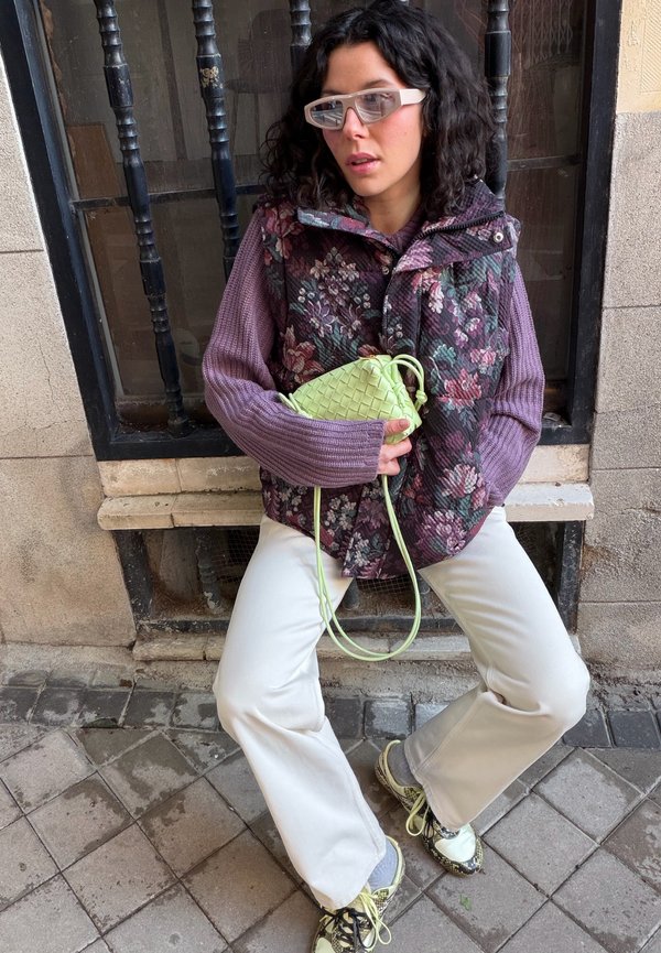 Donna con capelli ricci e scuri che indossa un gilet floreale e un maglione viola, che tiene in mano una piccola borsa tessuta di colore verde chiaro, seduta di fronte a una finestra con sbarre.